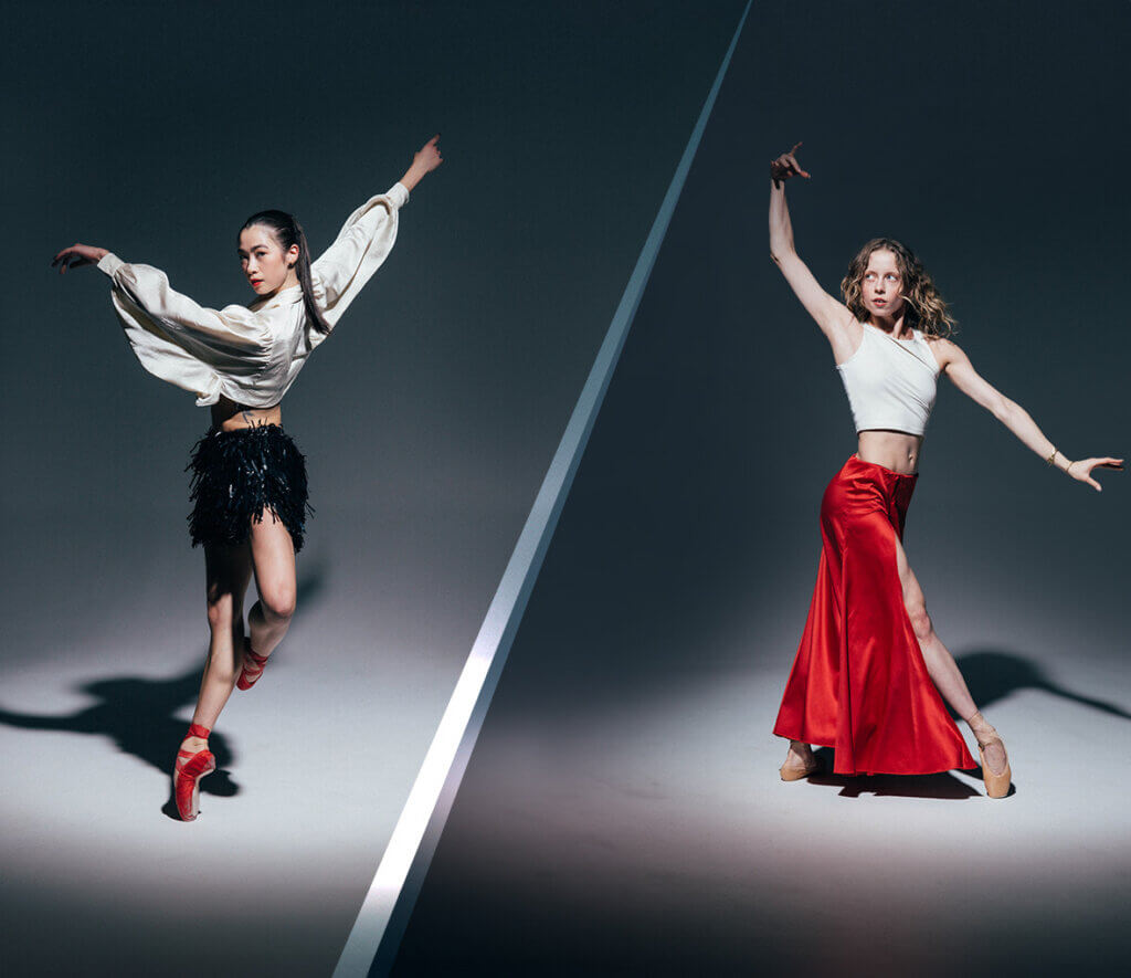 Two females dancing in moody lighting. Once dancer us wearing a flowy white crop top with long sleeves and black shorts with fringe and red pointe shoes. The other dancer is wearing a tight white tank top and a long red skirt with nude pointe shoes.