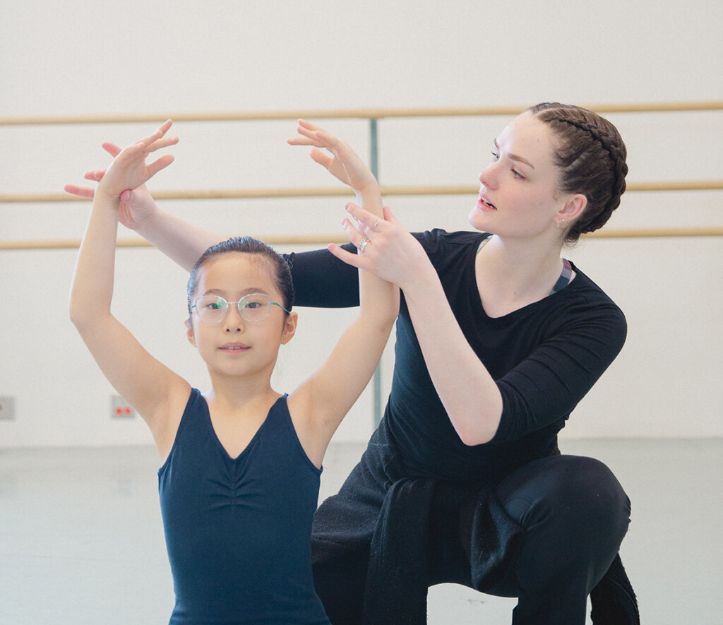a young Asian girl in class practising ballet with an instructor behind her correcting her arm placement