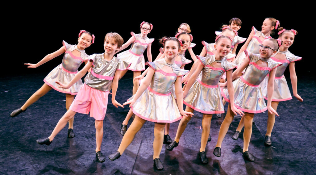De jeunes danseurs souriants se produisent sur scène dans des costumes spatiaux argentés et roses