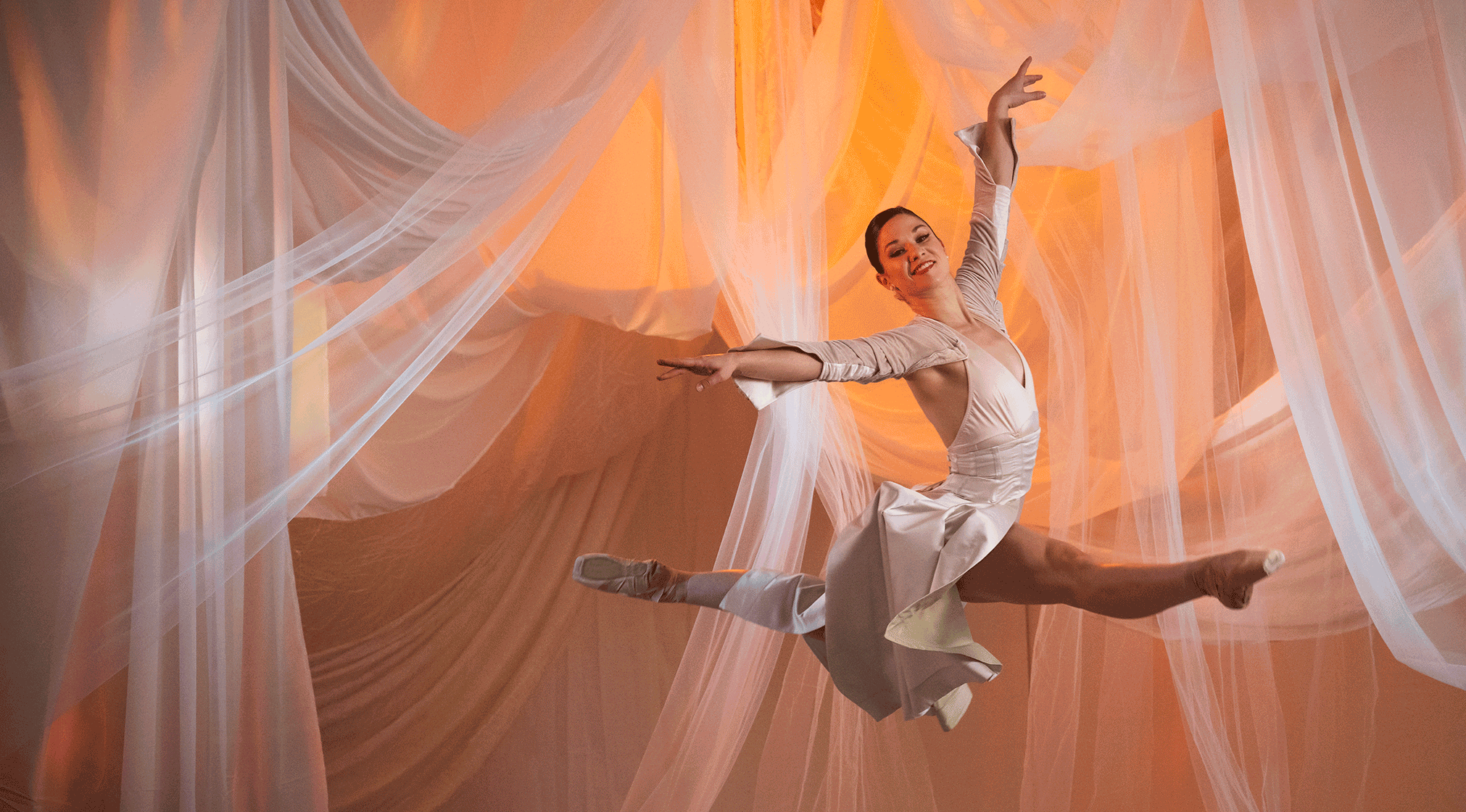 A female dancer in a jumping pose against a backdrop of hanging fabric in warm orange lighting.