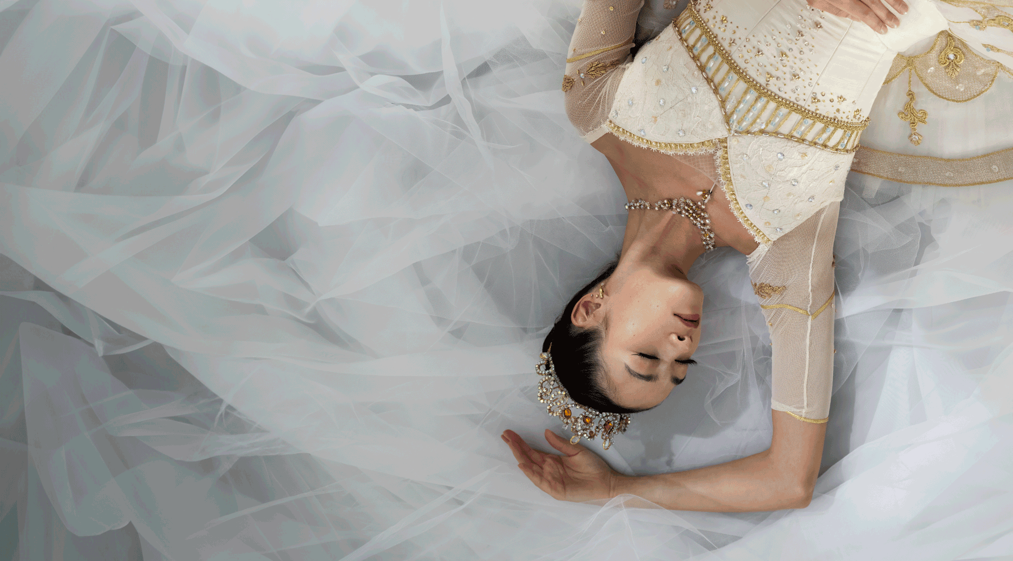 A female dancer dressed in a white and gold tutu and crown sleeping among white fabric